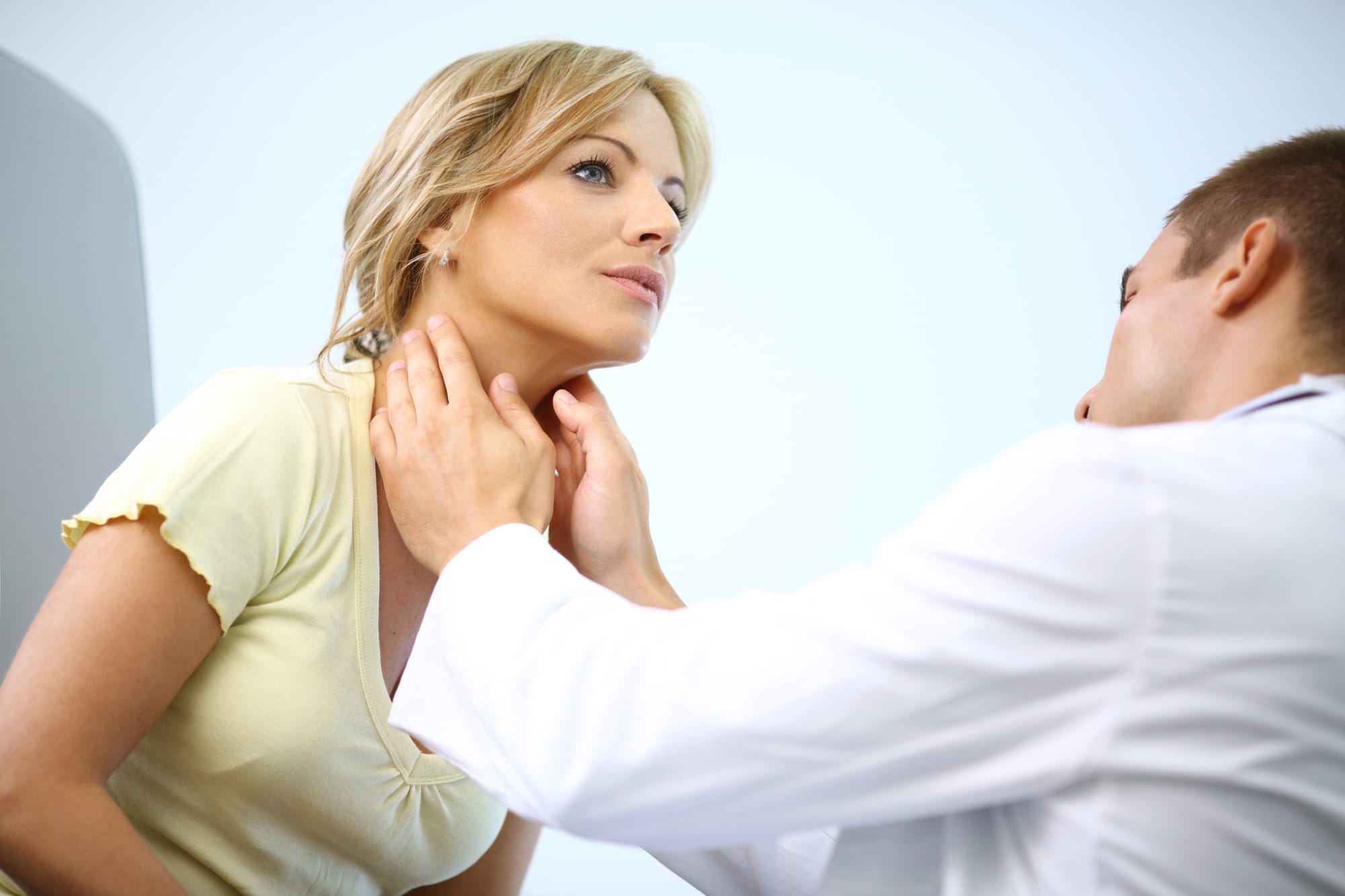 Middle aged blond woman visiting general practitoner.She's in examination room while doctor is touching her neck cause she has sore and swollen tonsils.She's looking up.